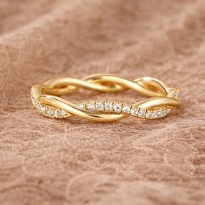 Può includere: Un anello dorato con un design intrecciato. L'anello è adornato con piccole pietre preziose trasparenti. L'anello è posto su uno sfondo neutro e strutturato.