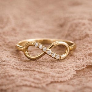 Puede incluir: Un anillo dorado con un diseño de símbolo de infinito. El símbolo del infinito está adornado con pequeñas gemas brillantes. El anillo se presenta sobre un fondo suave y texturizado, resaltando el elegante diseño de la joya.