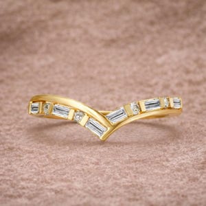 Pode incluir: Um anel de ouro em forma de V com diamantes baguette e redondos. O anel tem um acabamento polido e está sobre um fundo macio e neutro. Os diamantes são claros e brilhantes.