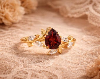 May include: A gold ring featuring a pear-shaped, deep red gemstone as the focal point. The ring's band and setting are adorned with delicate leaf and white gemstone accents, creating an elegant, nature-inspired design. The ring is set against a soft, textured background.