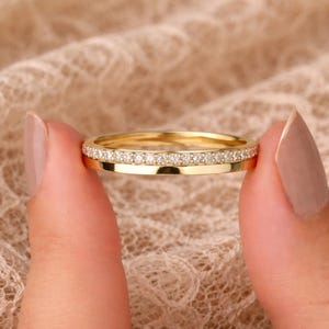 Op de afbeelding: Een gouden bandring met een rij kleine, heldere edelstenen langs de bovenrand. De ring wordt tussen twee vingers gehouden tegen een zachte, getextureerde achtergrond.