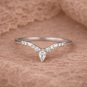 May include: A silver ring with a curved design, featuring a pear-shaped central stone and smaller round and baguette-cut stones. The ring is set against a soft, textured background, highlighting its elegant details.