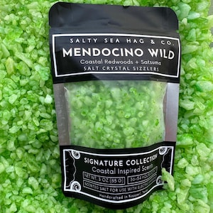 Op de afbeelding: Een zwart pakketje Mendocino Wild zoutkristal bruisballen, met de tekst "Salty Sea Hag & Co." en "Coastal Redwoods + Satsuma". Het pakketje bevat groene kristallen en is omringd door meer groene kristallen. Nettogewicht: 85 g.
