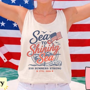 May include: An ivory tank top featuring a graphic print with the text Sea to Shining Sea, Sew Sling, and 250 Summers Strong 1776-2026. The design includes a compass rose, a small American flag, and birds. The garment is worn with a red, white, and blue hat.