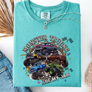 Peut inclure: T-shirt vert d'eau avec un motif de monster trucks. Le design pr&eacute;sente plusieurs camions de diff&eacute;rentes couleurs, avec le texte "Monster Trucks Are My Jam". Un collier argent&eacute; est drap&eacute; sur le t-shirt.