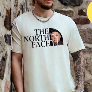 May include: Cream-colored t-shirt with the text "THE NORTH FACE" in black, featuring a parody image. The shirt has short sleeves and a crew neck. The image is of a person wearing the shirt.