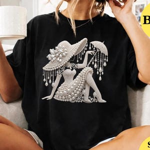 May include: Black t-shirt featuring a detailed illustration of a woman in a hat and dress adorned with pearls. The design includes an umbrella and is rendered in shades of white and silver. The shirt is a classic crew neck style.