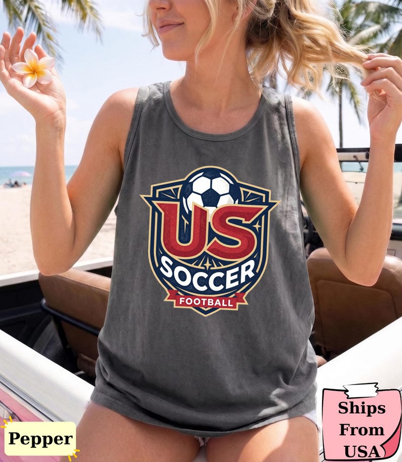 May include: A charcoal gray sleeveless tank top featuring a graphic design on the chest. The design includes a soccer ball above the red text US, the white text SOCCER, and the red banner with the white text FOOTBALL, all inside a shield emblem.