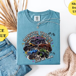 Peut inclure: T-shirt bleu glacier avec un motif de plusieurs monster trucks de diff&eacute;rentes couleurs. Le texte "Monster Trucks Are My Jam" est imprim&eacute; sur le t-shirt. Un collier argent&eacute; avec un pendentif est sur le t-shirt. Le t-shirt est pli&eacute; sur une surface blanche.