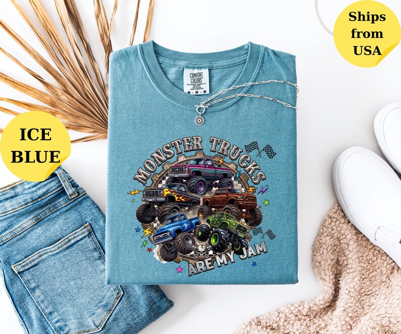 Peut inclure: T-shirt bleu glacier avec un motif de plusieurs monster trucks de diff&eacute;rentes couleurs. Le texte "Monster Trucks Are My Jam" est imprim&eacute; sur le t-shirt. Un collier argent&eacute; avec un pendentif est sur le t-shirt. Le t-shirt est pli&eacute; sur une surface blanche.