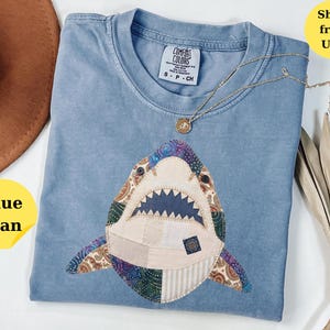 May include: A blue jean colored t-shirt featuring a large, central shark applique made from various patterned fabric patches. The shark design includes dark blue teeth, button eyes, and visible stitching. A thin gold chain necklace rests on the shirt.