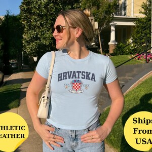 May include: A light blue short-sleeved t-shirt featuring the text HRVATSKA in a dark blue arched font above a crest graphic with a red and white checkered pattern, small gold stars, and the text EST. 1991 on a white banner.
