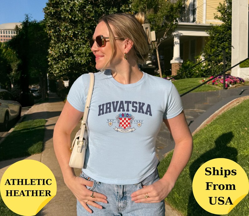 May include: A light blue short-sleeved t-shirt featuring the text HRVATSKA in a dark blue arched font above a crest graphic with a red and white checkered pattern, small gold stars, and the text EST. 1991 on a white banner.
