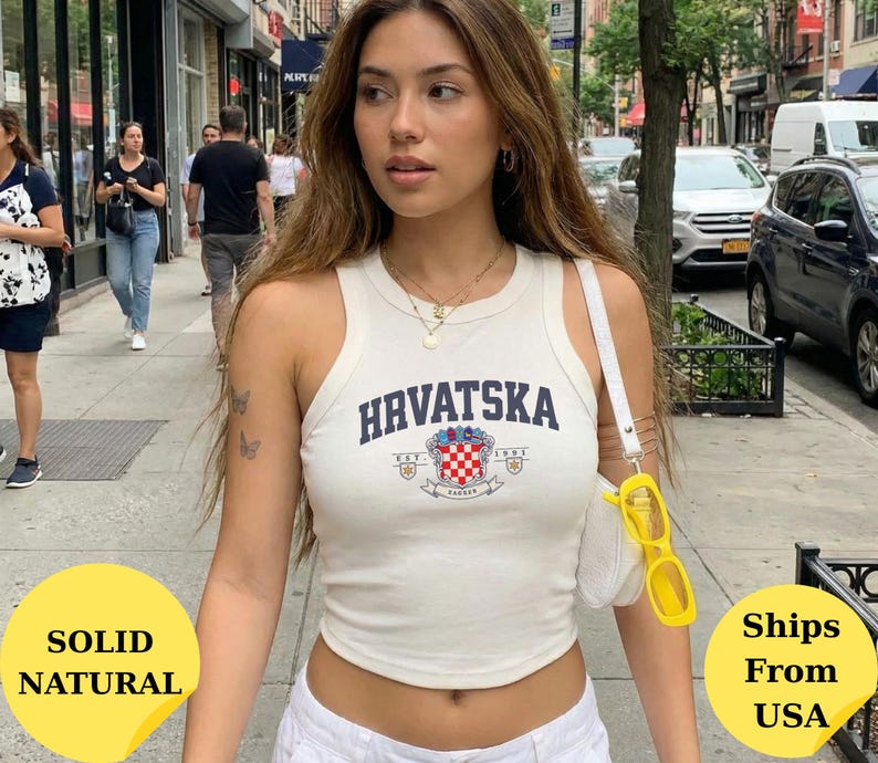 May include: A white sleeveless tank top featuring the text HRVATSKA in dark blue letters above a colorful crest emblem with a red and white checkered pattern. The top has a round neckline and a fitted silhouette, worn with a white shoulder bag.