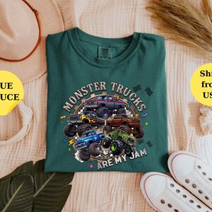 Peut inclure: Un t-shirt vert d'eau avec un motif de plusieurs monster trucks de diff&eacute;rentes couleurs, dont le violet, le bleu et le vert. Le texte "MONSTER TRUCKS ARE MY JAM" est imprim&eacute; autour des camions. Le t-shirt est pr&eacute;sent&eacute; sur une surface claire.