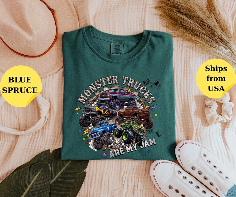 Peut inclure: Un t-shirt vert d'eau avec un motif de plusieurs monster trucks de diff&eacute;rentes couleurs, dont le violet, le bleu et le vert. Le texte "MONSTER TRUCKS ARE MY JAM" est imprim&eacute; autour des camions. Le t-shirt est pr&eacute;sent&eacute; sur une surface claire.