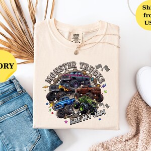 Peut inclure: T-shirt ivoire avec un motif de plusieurs monster trucks de diff&eacute;rentes couleurs. Le texte "MONSTER TRUCKS ARE MY JAM" est imprim&eacute; dans un design circulaire. Le t-shirt est pos&eacute; &agrave; plat avec un jean bleu et des chaussures blanches.