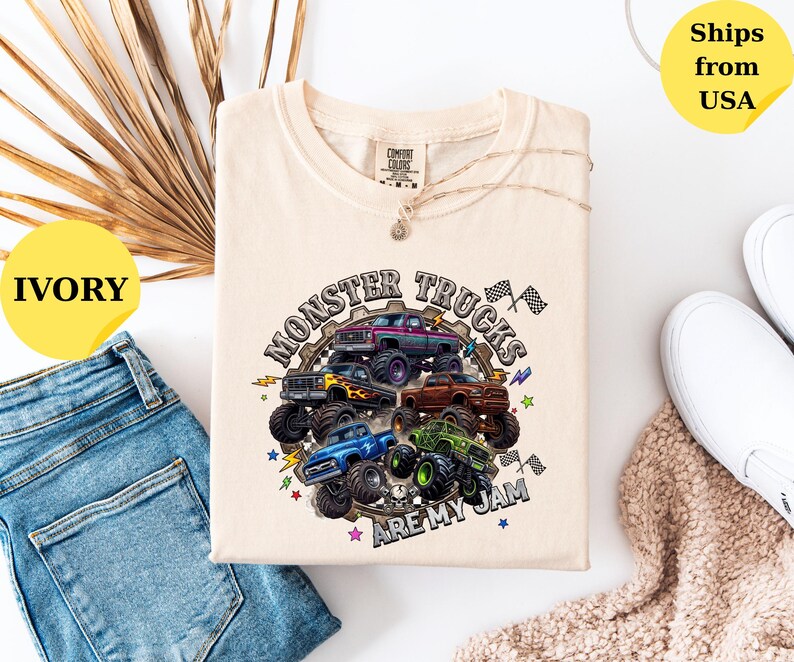 Peut inclure: T-shirt ivoire avec un motif de plusieurs monster trucks de diff&eacute;rentes couleurs. Le texte "MONSTER TRUCKS ARE MY JAM" est imprim&eacute; dans un design circulaire. Le t-shirt est pos&eacute; &agrave; plat avec un jean bleu et des chaussures blanches.
