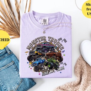 Peut inclure: T-shirt de couleur orchid&eacute;e avec un motif de monster trucks. Le design pr&eacute;sente plusieurs camions de diff&eacute;rentes couleurs, avec le texte "MONSTER TRUCKS ARE MY JAM". Le t-shirt est pr&eacute;sent&eacute; avec un jean bleu et des chaussures blanches.