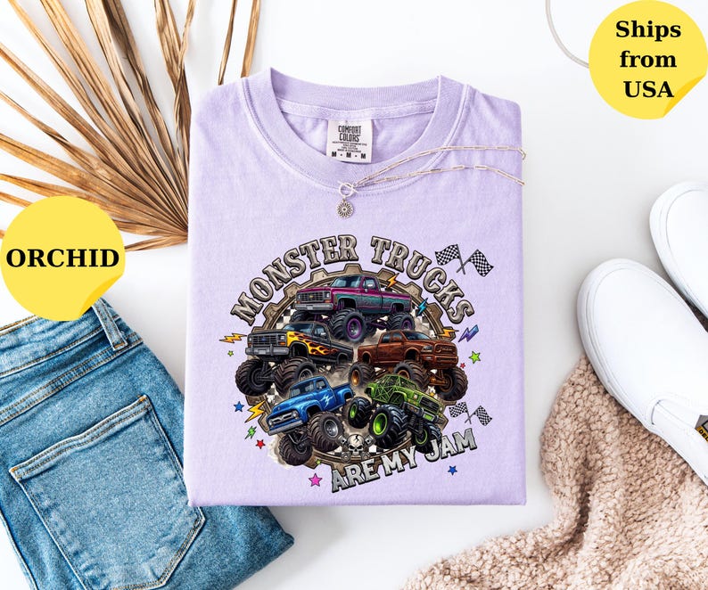 Peut inclure: T-shirt de couleur orchid&eacute;e avec un motif de monster trucks. Le design pr&eacute;sente plusieurs camions de diff&eacute;rentes couleurs, avec le texte "MONSTER TRUCKS ARE MY JAM". Le t-shirt est pr&eacute;sent&eacute; avec un jean bleu et des chaussures blanches.