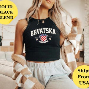 May include: A black sleeveless tank top featuring the white text HRVATSKA in an arched font above a graphic of the Croatian coat of arms with the text EST. 1991. The garment is worn with a beige and white checkered cardigan and light gray sweatpants.