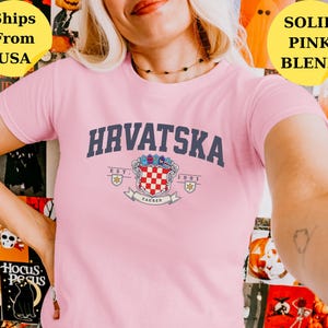 May include: A light pink short-sleeve t-shirt featuring the dark blue text HRVATSKA arched above a colorful coat of arms emblem. The emblem includes a red and white checkered shield, the text EST. 1991, and a banner with the word ZAGREB.
