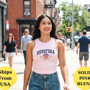May include: A light pink sleeveless tank top featuring the text HRVATSKA in dark blue letters above a crest with a red and white checkered pattern. The garment has a ribbed texture and a high neckline, paired with blue denim jeans.