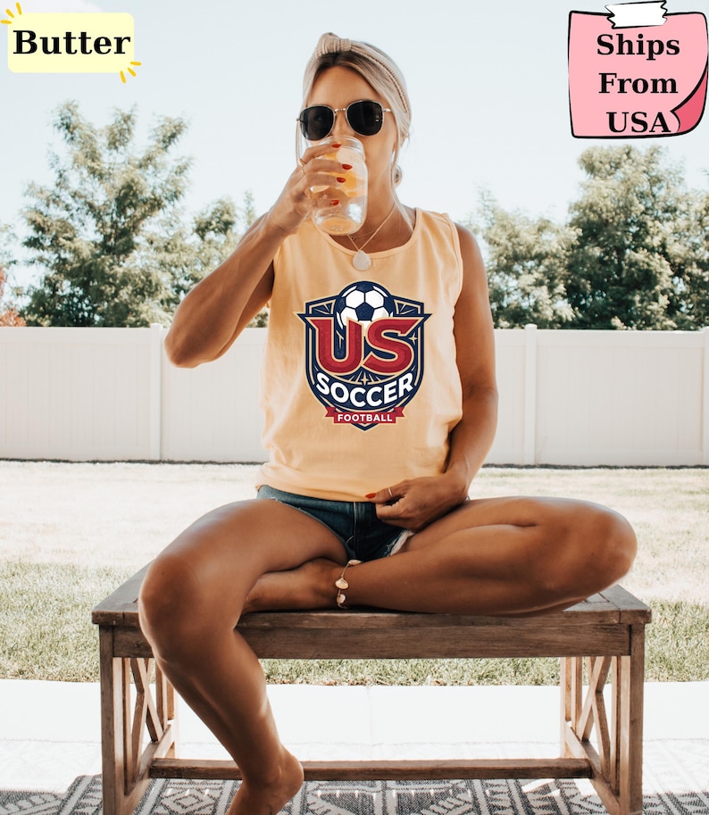 May include: A sleeveless butter yellow tank top featuring a graphic logo on the chest with a soccer ball, the text US SOCCER, and the word FOOTBALL in a red banner. The design includes navy blue and red accents on a light yellow background.