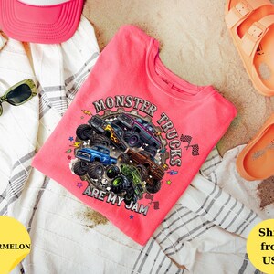 Peut inclure: Un t-shirt rose avec un motif de monster trucks et le texte "MONSTER TRUCKS ARE MY JAM". L'image comprend &eacute;galement une casquette de camionneur rose, des lunettes de soleil et des sandales orange. Le t-shirt est pr&eacute;sent&eacute; sur un tissu &agrave; rayures blanches et grises.