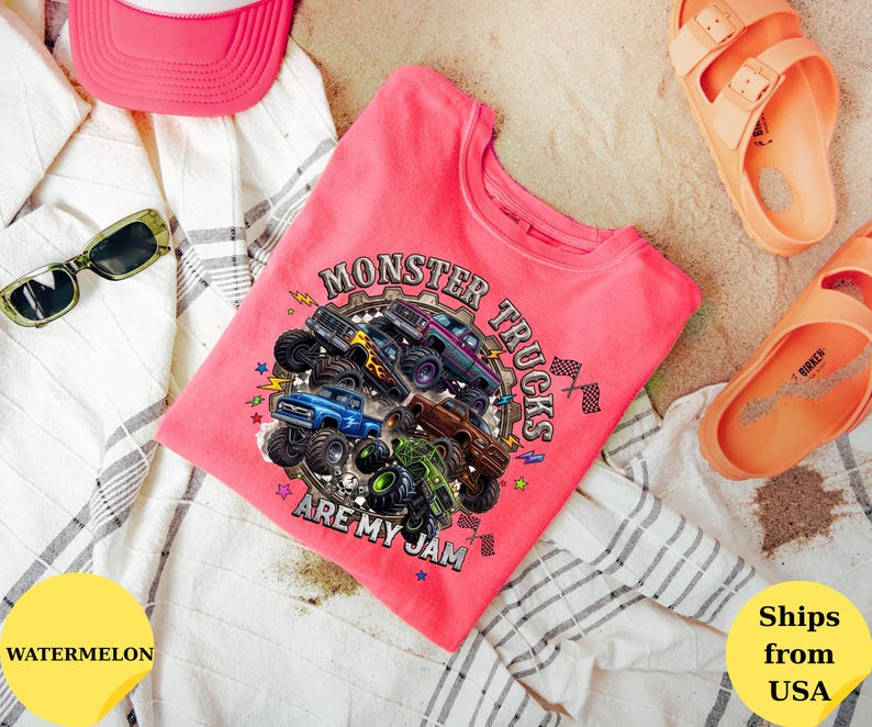 Peut inclure: Un t-shirt rose avec un motif de monster trucks et le texte "MONSTER TRUCKS ARE MY JAM". L'image comprend &eacute;galement une casquette de camionneur rose, des lunettes de soleil et des sandales orange. Le t-shirt est pr&eacute;sent&eacute; sur un tissu &agrave; rayures blanches et grises.
