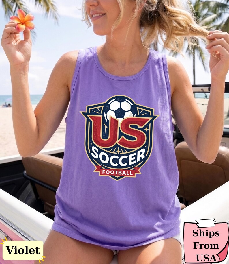 May include: A violet sleeveless tank top featuring a graphic print of a soccer ball above the text US SOCCER and FOOTBALL in a shield-shaped emblem with red, dark blue, and white colors. The design is centered on the front of the garment.