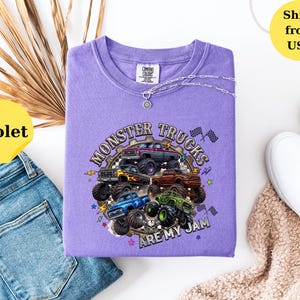 Peut inclure: Un t-shirt violet Comfort Colors avec un motif de monster trucks et le texte "MONSTER TRUCKS ARE MY JAM". Un collier argent&eacute; est drap&eacute; sur le t-shirt. Le t-shirt est pr&eacute;sent&eacute; avec un jean bleu et des chaussures blanches. Le t-shirt est d'une nuance de violet vif.