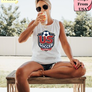 May include: A white sleeveless tank top featuring a graphic logo on the chest. The logo displays a soccer ball above the red text US and the blue text SOCCER, with the word FOOTBALL on a red banner at the bottom, all enclosed in a shield design.