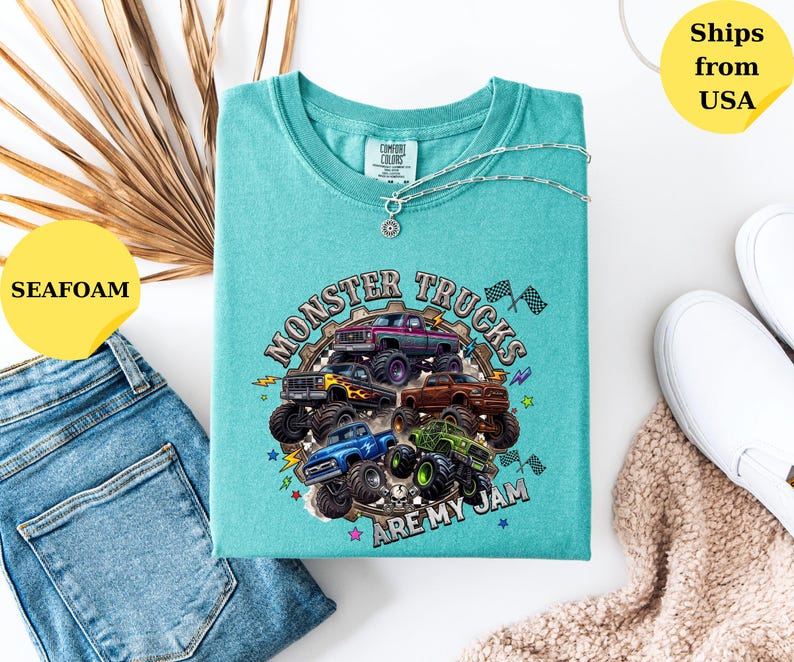 Peut inclure: T-shirt vert d'eau avec un motif de monster trucks. Le design pr&eacute;sente plusieurs camions de diff&eacute;rentes couleurs, avec le texte "Monster Trucks Are My Jam". Un collier argent&eacute; est drap&eacute; sur le t-shirt.