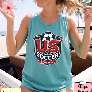May include: A seafoam green sleeveless tank top featuring a graphic design of a soccer ball above the text US SOCCER and FOOTBALL in a shield-shaped emblem with red, white, and blue colors. The design is centered on the front of the garment.