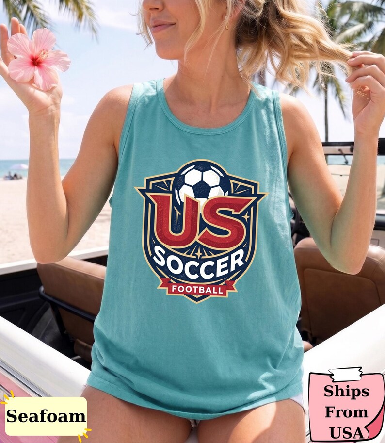 May include: A seafoam green sleeveless tank top featuring a graphic design of a soccer ball above the text US SOCCER and FOOTBALL in a shield-shaped emblem with red, white, and blue colors. The design is centered on the front of the garment.