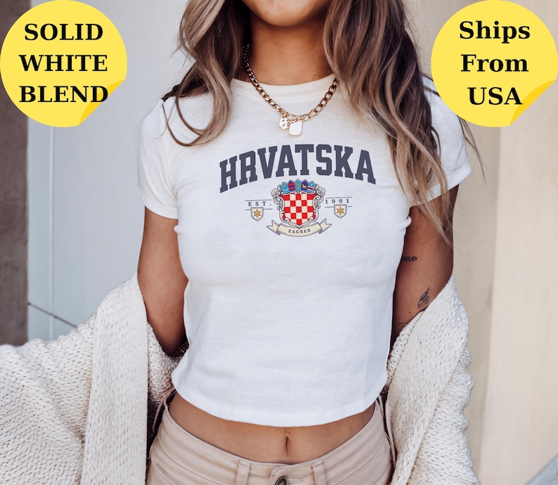 May include: A white short-sleeved t-shirt featuring the text HRVATSKA in dark blue letters above a colorful crest graphic with the text EST. 1991 and ZAGREB. The shirt has a crew neckline and is worn with a gold-toned chain necklace and a beige cardigan.