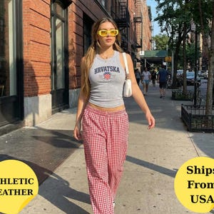 May include: A grey sleeveless tank top featuring the text HRVATSKA in dark blue letters above a small red and white checkered shield emblem, paired with red and white gingham patterned trousers and a small white shoulder bag with yellow sunglasses.