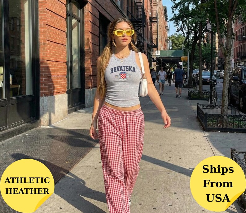 May include: A grey sleeveless tank top featuring the text HRVATSKA in dark blue letters above a small red and white checkered shield emblem, paired with red and white gingham patterned trousers and a small white shoulder bag with yellow sunglasses.