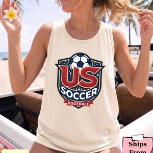 May include: An ivory sleeveless tank top featuring a graphic print of a soccer ball inside a shield emblem. The design includes the red text US, the white text SOCCER, and the red text FOOTBALL on a banner, all set against a dark blue background.