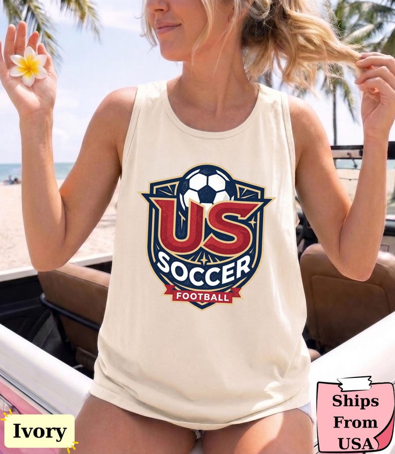 May include: An ivory sleeveless tank top featuring a graphic print of a soccer ball inside a shield emblem. The design includes the red text US, the white text SOCCER, and the red text FOOTBALL on a banner, all set against a dark blue background.