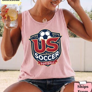 May include: A light pink sleeveless tank top featuring a graphic print of a soccer ball inside a shield emblem with the text US SOCCER FOOTBALL in red, white, and dark blue colors. The design is centered on the chest of the garment.