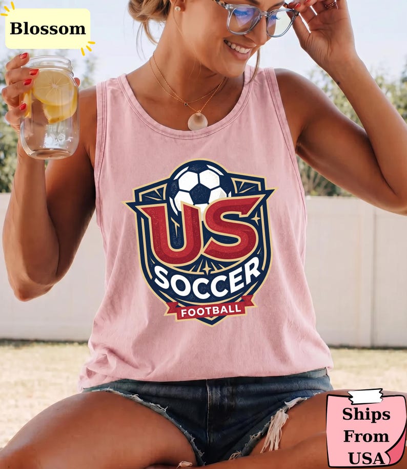 May include: A light pink sleeveless tank top featuring a graphic print of a soccer ball inside a shield emblem with the text US SOCCER FOOTBALL in red, white, and dark blue colors. The design is centered on the chest of the garment.