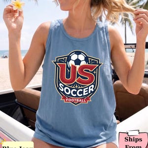 May include: A blue sleeveless tank top featuring a graphic emblem on the chest. The design includes a soccer ball above large red letters reading US, with the word SOCCER in white below, and a red banner at the bottom with the word FOOTBALL.