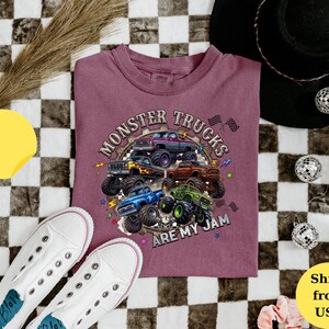 Peut inclure: T-shirt bordeaux avec un motif de plusieurs monster trucks color&eacute;s. Le texte "MONSTER TRUCKS ARE MY JAM" est imprim&eacute; au-dessus et en dessous des camions. Des baskets blanches sont visibles en bas &agrave; gauche.