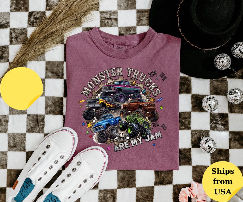 Peut inclure: T-shirt bordeaux avec un motif de plusieurs monster trucks color&eacute;s. Le texte "MONSTER TRUCKS ARE MY JAM" est imprim&eacute; au-dessus et en dessous des camions. Des baskets blanches sont visibles en bas &agrave; gauche.
