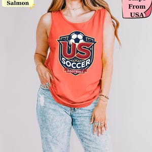 May include: A bright salmon sleeveless tank top featuring a graphic print of a soccer ball above the text US SOCCER FOOTBALL. The design is enclosed in a dark blue and gold shield emblem with decorative star accents and a banner at the bottom.