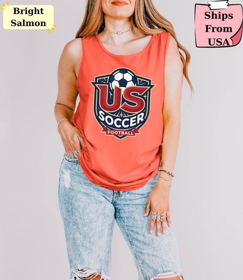 May include: A bright salmon sleeveless tank top featuring a graphic print of a soccer ball above the text US SOCCER FOOTBALL. The design is enclosed in a dark blue and gold shield emblem with decorative star accents and a banner at the bottom.