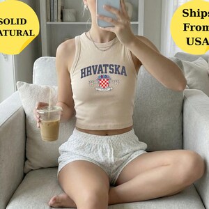 May include: A beige ribbed tank top featuring the text HRVATSKA in dark blue letters above a colorful Croatian coat of arms graphic. The sleeveless top has a high crew neckline and a cropped fit, paired with light-colored patterned shorts.