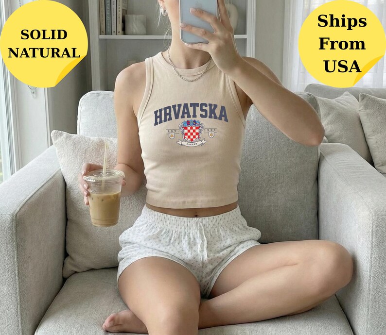 May include: A beige ribbed tank top featuring the text HRVATSKA in dark blue letters above a colorful Croatian coat of arms graphic. The sleeveless top has a high crew neckline and a cropped fit, paired with light-colored patterned shorts.